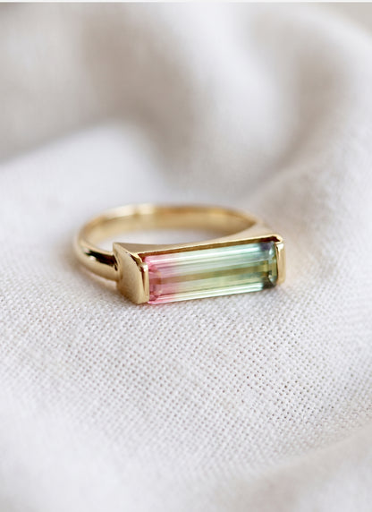 A gold ring with a rectangular baguette-cut iridescent gemstone showing pink, green, and blue color shifts, resting on a textured white fabric background.