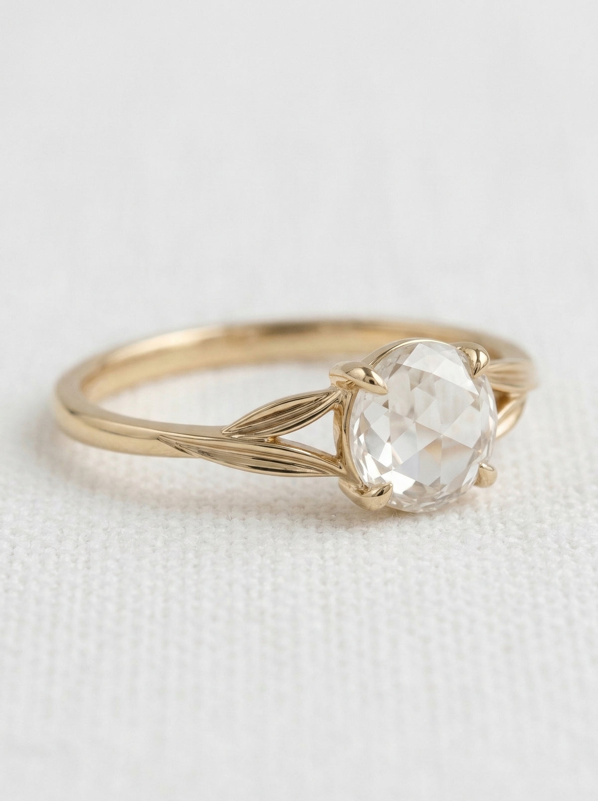 A delicate gold ring featuring a large, rose-cut clear diamond in a prong setting, resting on a textured white fabric background.