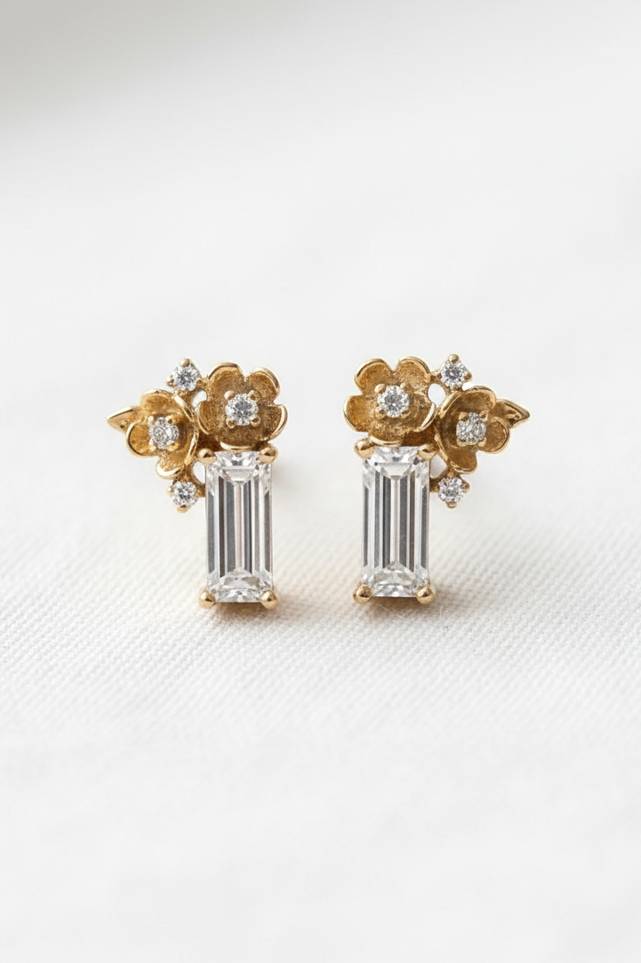 A pair of gold and white gold stud earrings featuring a floral design with clear baguette and round-cut stones, displayed on a clean white textured surface.