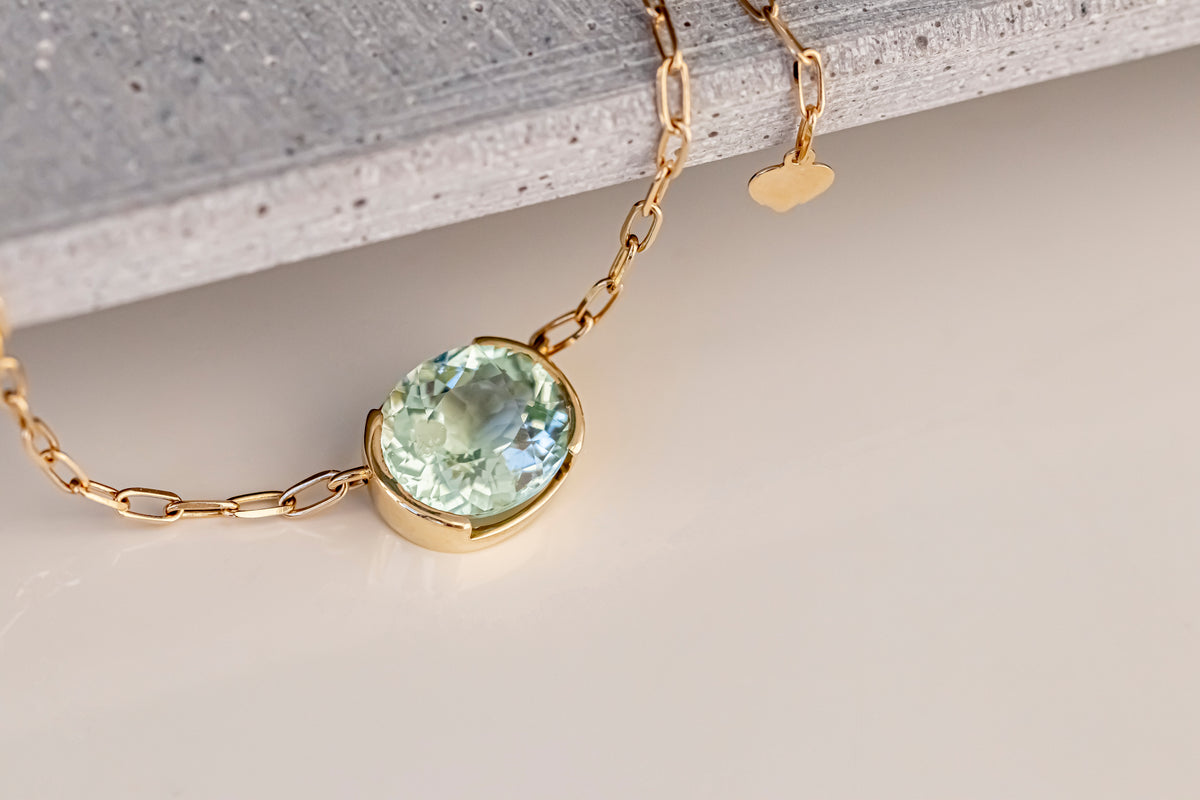 A gold chain necklace with a bezel-set oval light green gemstone pendant, displayed on a neutral light beige surface with a textured gray stone prop.