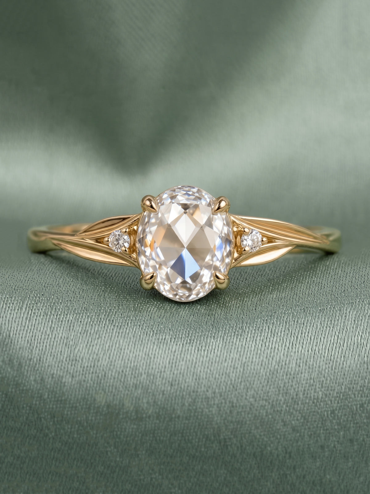 A yellow gold engagement ring featuring a large oval-cut clear center stone and two small round side stones, displayed on a textured sage green fabric background.