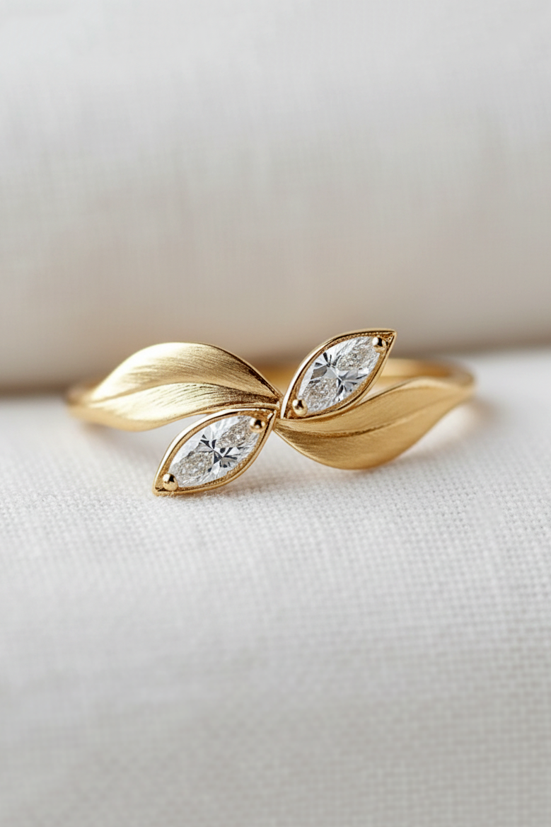 A gold ring designed like two overlapping leaves, each with a small marquise-cut clear gemstone, resting on a textured white fabric surface.
