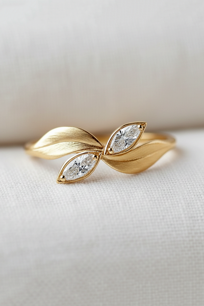 A gold ring designed like two overlapping leaves, each with a small marquise-cut clear gemstone, resting on a textured white fabric surface.