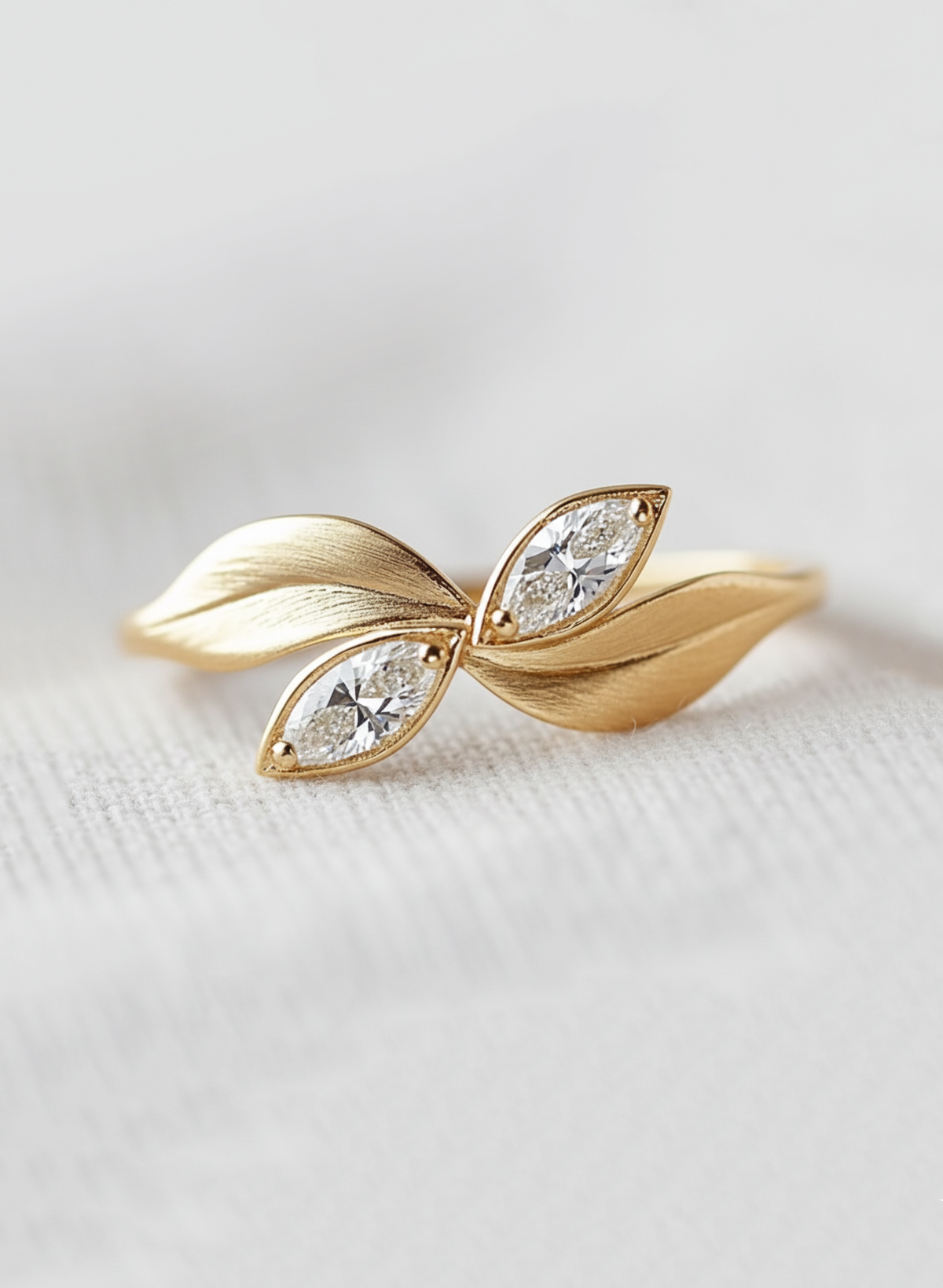 A gold leaf-shaped ring featuring two marquise-cut clear gemstones, displayed on a soft, light gray textured fabric background.