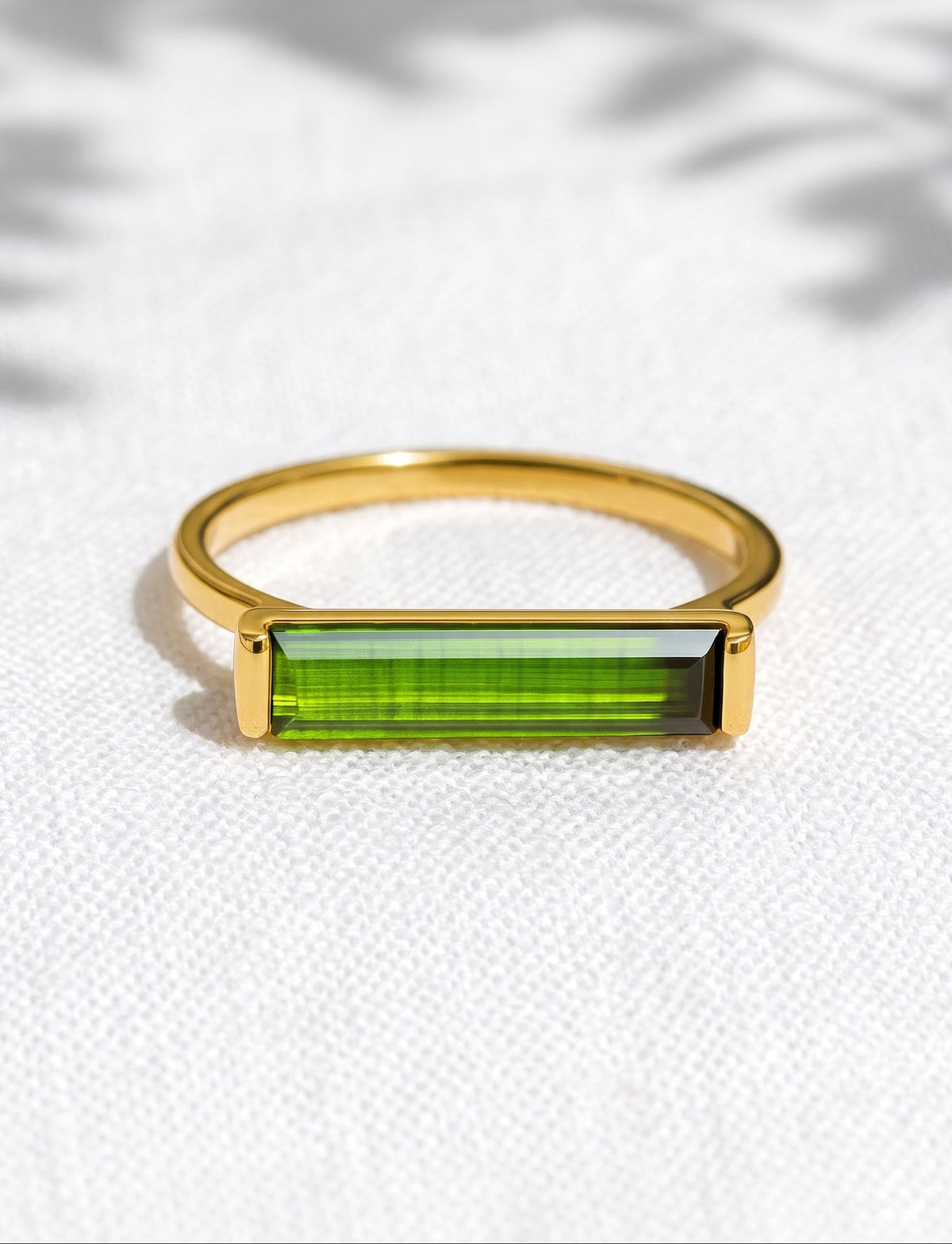 A gold ring with a rectangular green gemstone, resting on a textured white fabric background with soft leaf shadows.