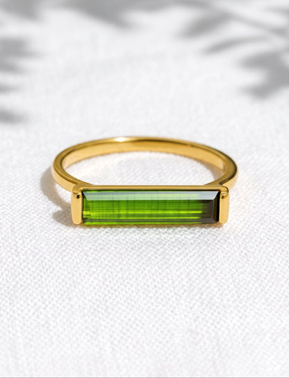 A gold ring with a rectangular green gemstone, resting on a textured white fabric background with soft leaf shadows.
