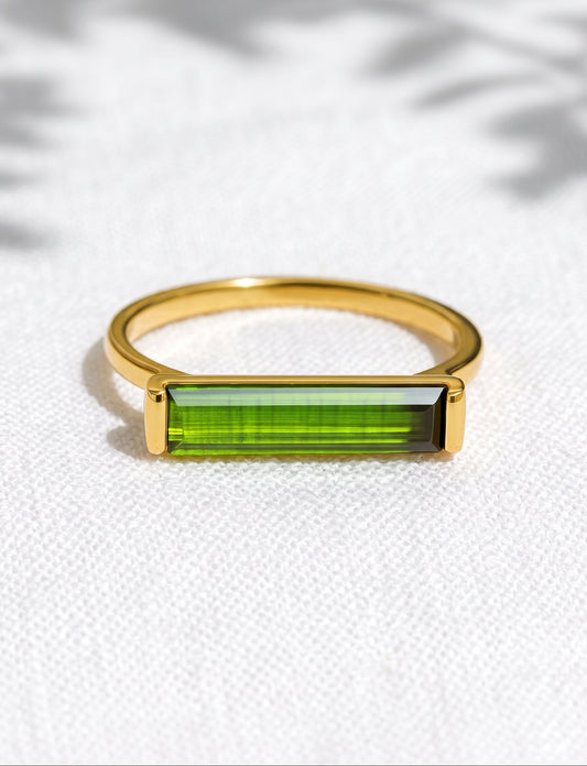 A gold ring with a rectangular green gemstone, resting on a textured white fabric background with soft leaf shadows.