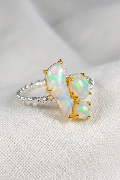 A close-up of a ring featuring three iridescent white opal stones in gold settings on a textured silver band, resting on a soft white fabric background.