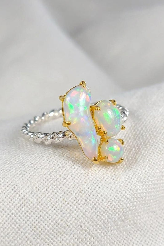 A close-up of a ring featuring three iridescent white opal stones in gold settings on a textured silver band, resting on a soft white fabric background.