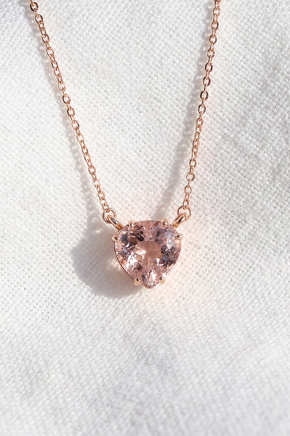 A delicate rose gold chain necklace featuring a small, heart-shaped pink gemstone pendant, displayed on a textured white fabric background.