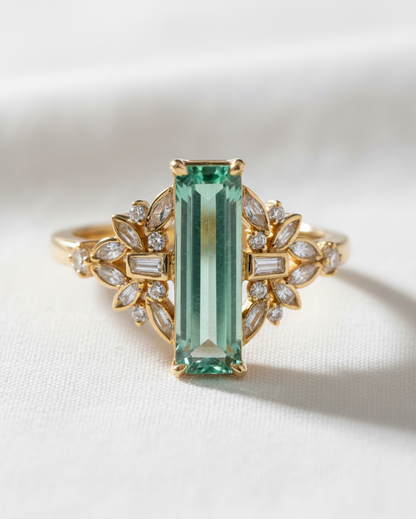 A yellow gold ring featuring a large rectangular emerald-cut green gemstone in the center, flanked by symmetrical leaf-shaped clusters of small clear baguette and round stones, set against a soft white fabric background.