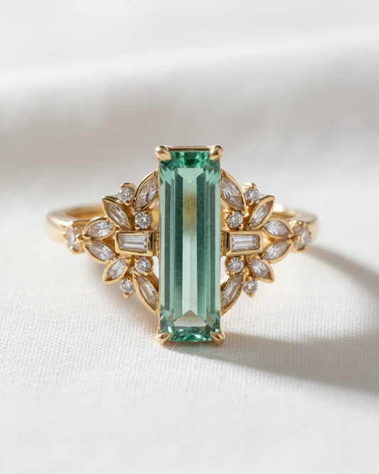 A yellow gold ring featuring a large rectangular emerald-cut green gemstone in the center, flanked by symmetrical leaf-shaped clusters of small clear baguette and round stones, set against a soft white fabric background.