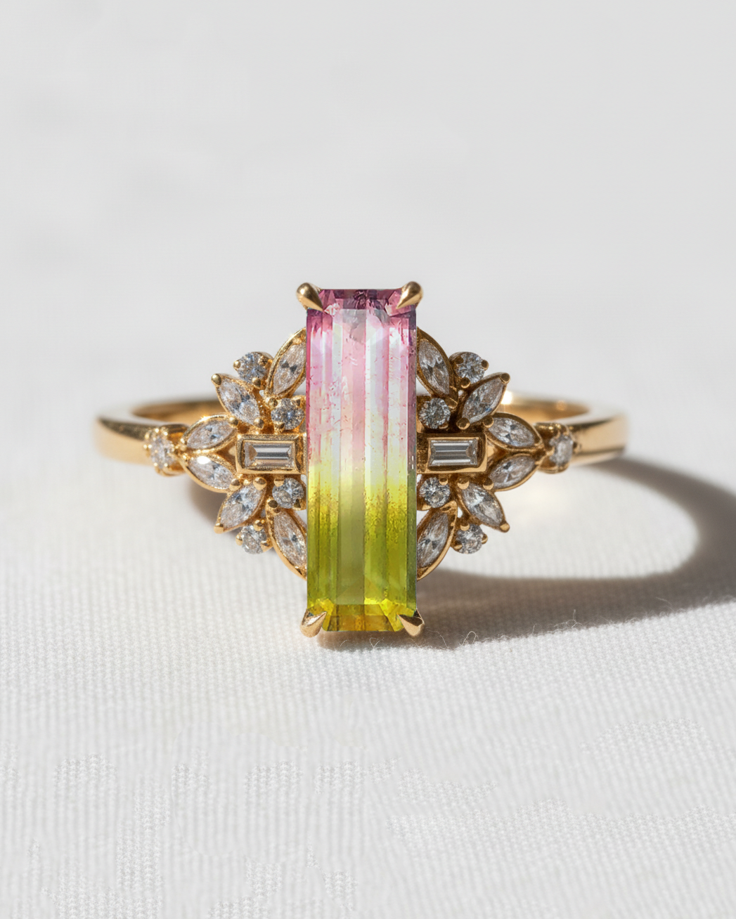 A yellow gold ring featuring a rectangular gradient tourmaline center stone that transitions from pink to green, accented by small clear stones on a white textured background.