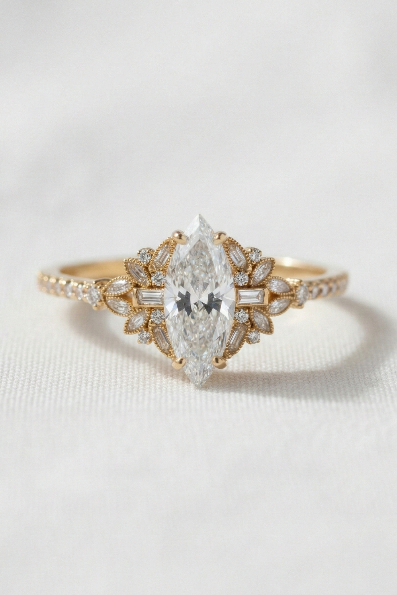 A yellow gold ring featuring a marquise-cut diamond center stone and smaller diamonds along the shoulders, set against a soft white textured background.