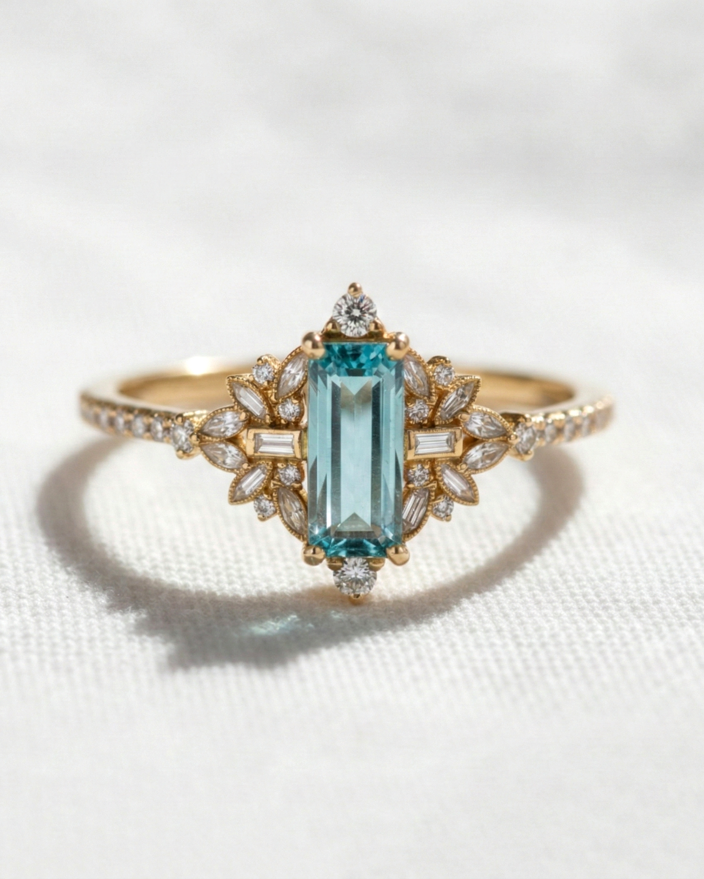 A yellow gold ring featuring a central emerald-cut light blue gemstone, surrounded by small clear accent stones in a floral setting, displayed on a textured white fabric background.