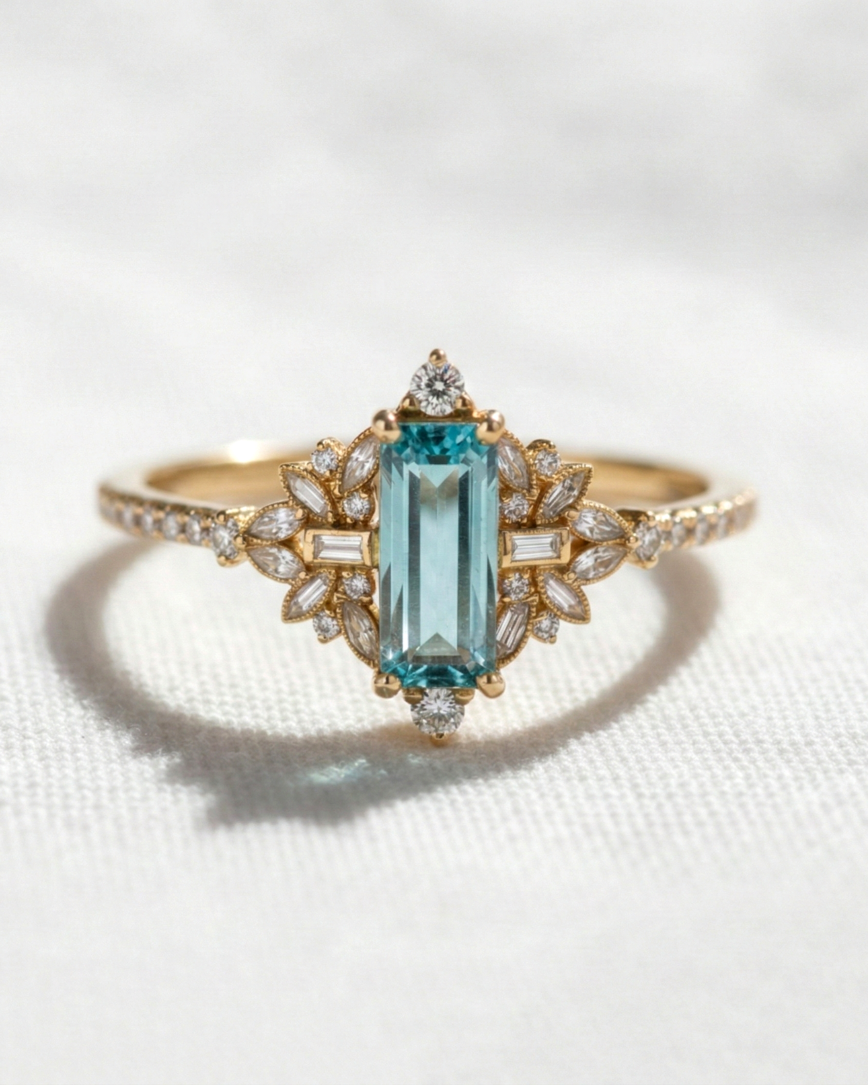 A yellow gold ring featuring a central emerald-cut light blue gemstone, surrounded by small clear accent stones in a floral setting, displayed on a textured white fabric background.
