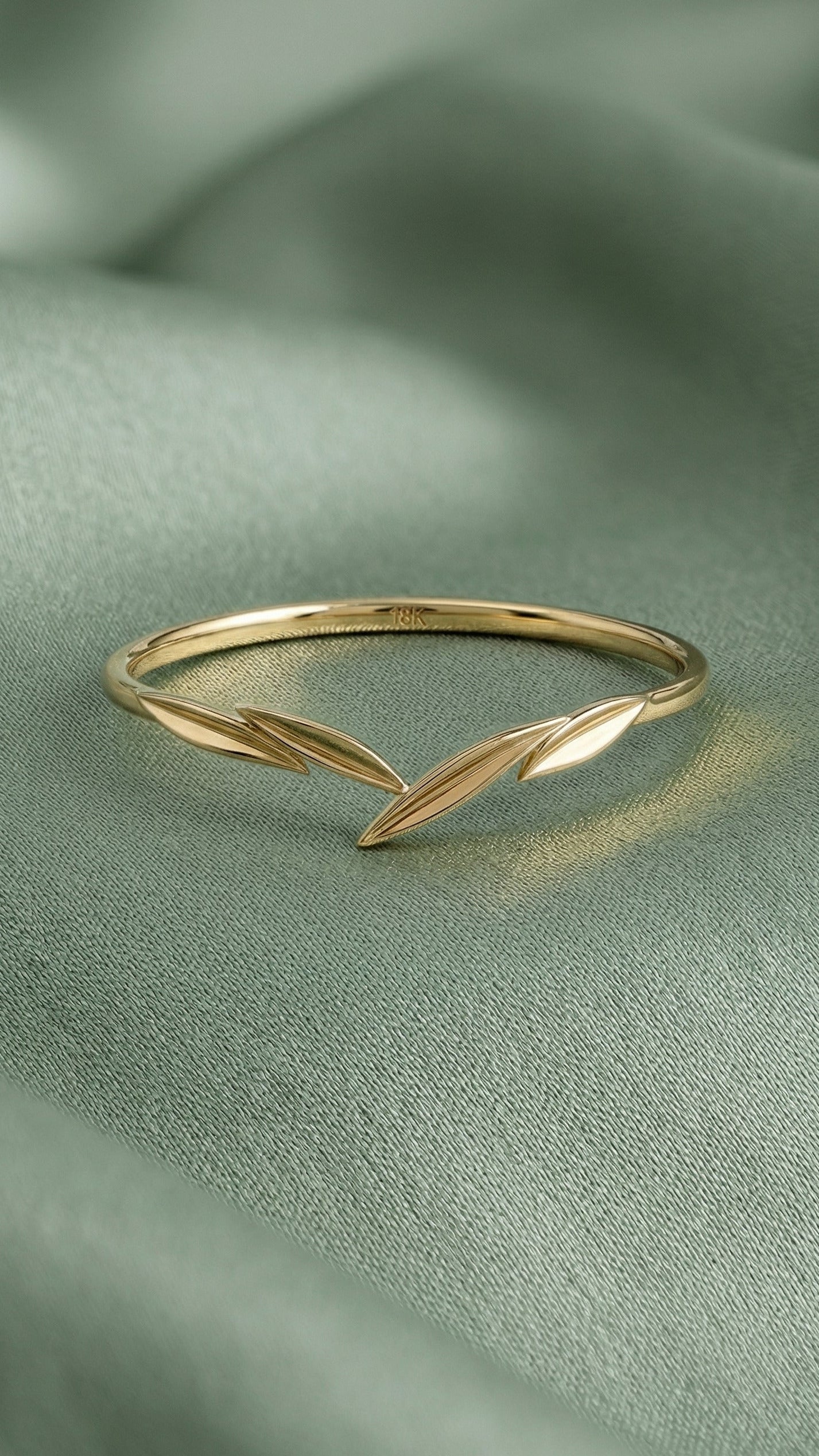 A delicate gold ring with a leaf design, resting on a soft, draped sage green fabric background.