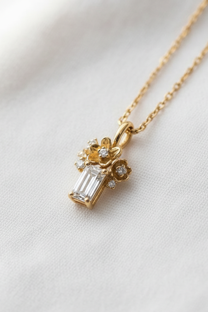 A delicate gold necklace with a pendant featuring a baguette-cut clear gemstone and small floral details, displayed on a soft white fabric background.