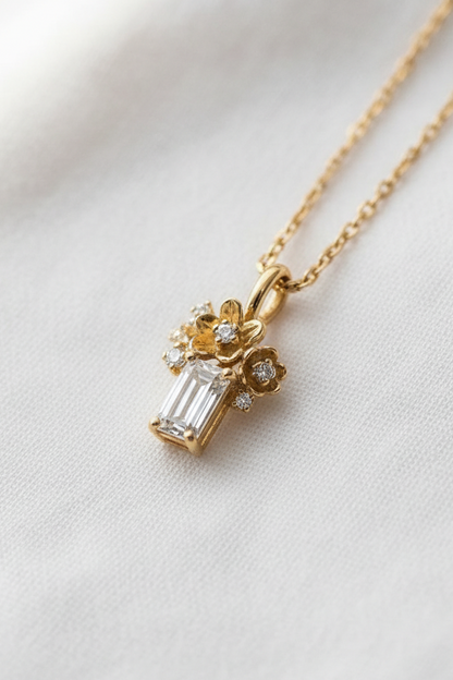 A delicate gold necklace with a pendant featuring a baguette-cut clear gemstone and small floral details, displayed on a soft white fabric background.