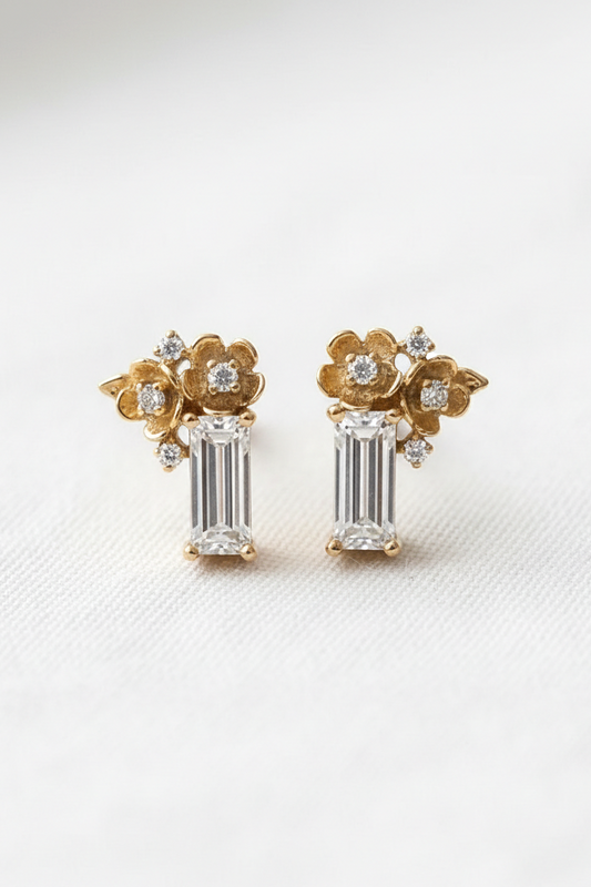 A pair of gold and white gold stud earrings featuring a floral design with clear baguette and round-cut stones, displayed on a clean white textured surface.