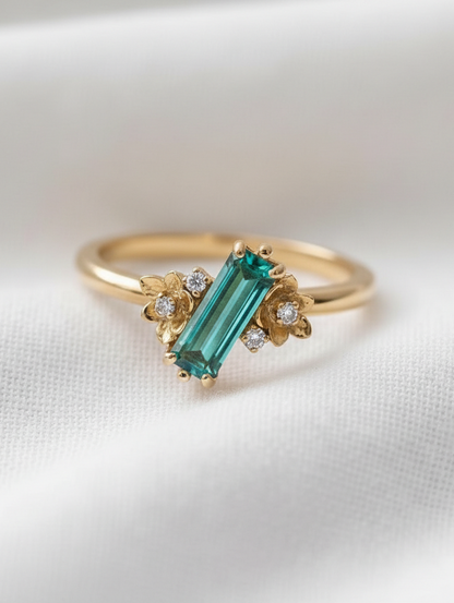 A gold ring featuring a rectangular baguette blue tourmaline gemstone, accented by small diamond-like stones in floral settings, resting on a white textured fabric background.
