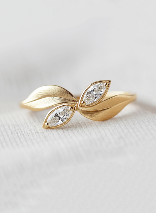 A gold leaf-shaped ring featuring two marquise-cut clear gemstones, displayed on a soft, light gray textured fabric background.
