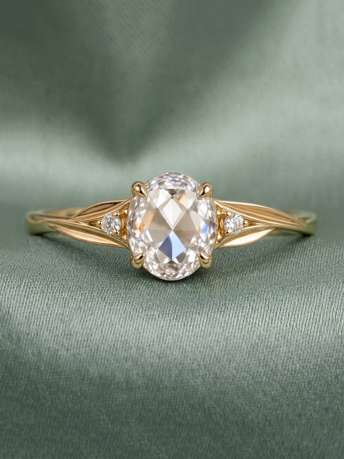 A yellow gold engagement ring featuring a large oval-cut clear center stone and two small round side stones, displayed on a textured sage green fabric background.