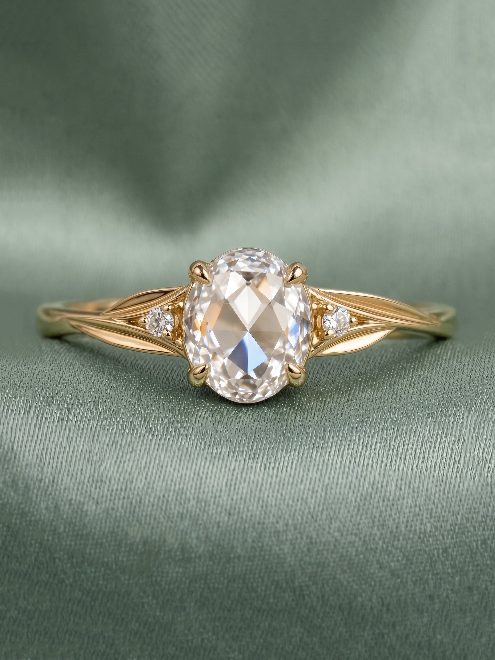 A yellow gold engagement ring featuring a large oval-cut clear center stone and two small round side stones, displayed on a textured sage green fabric background.