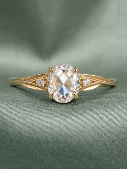 A yellow gold engagement ring featuring a large oval-cut clear center stone and two small round side stones, displayed on a textured sage green fabric background.