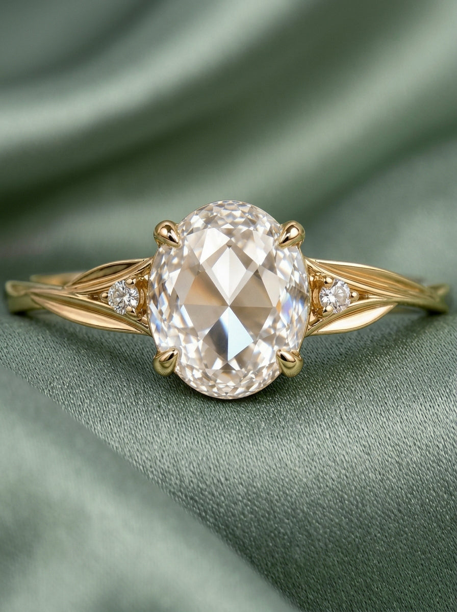 An oval-cut diamond engagement ring with a yellow gold band, resting on a draped sage green fabric background.