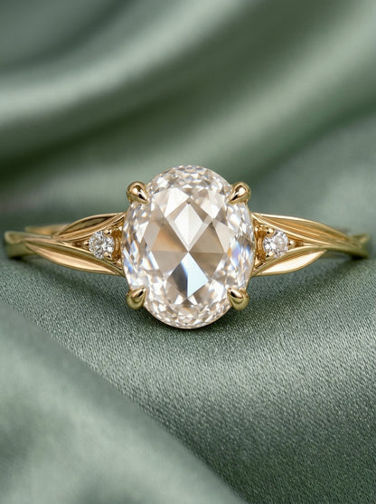 An oval-cut diamond engagement ring with a yellow gold band, resting on a draped sage green fabric background.