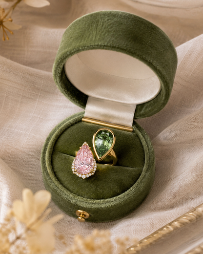 Two teardrop-shaped diamonds set in 18K gold displayed in an open olive green velvet ring box. One ring has a pink gemstone surrounded by small clear diamonds with a green diamond in bezel setting.